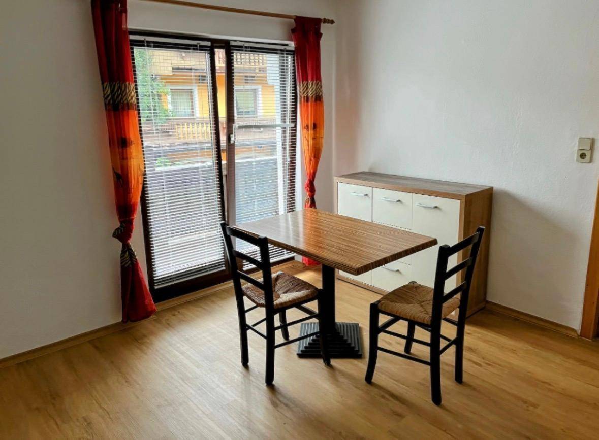 Essbereich mit Holztisch, zwei Stühlen und einem Holzschrank, beleuchtet durch ein Fenster mit roten Vorhängen.