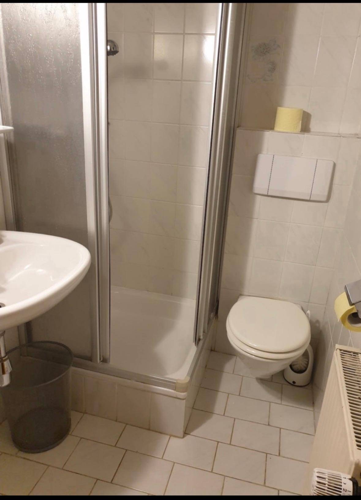 Kleines Badezimmer mit weißen Fliesen, Duschkabine, Toilette, Waschbecken und Heizkörper.