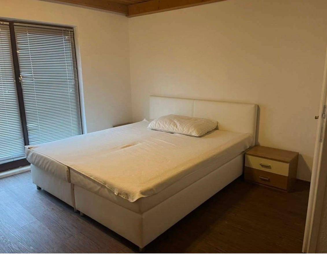 Schlafzimmer mit weißem Bett, Nachttisch und Fenster mit Jalousien, ergänzt durch eine Holzdecke.