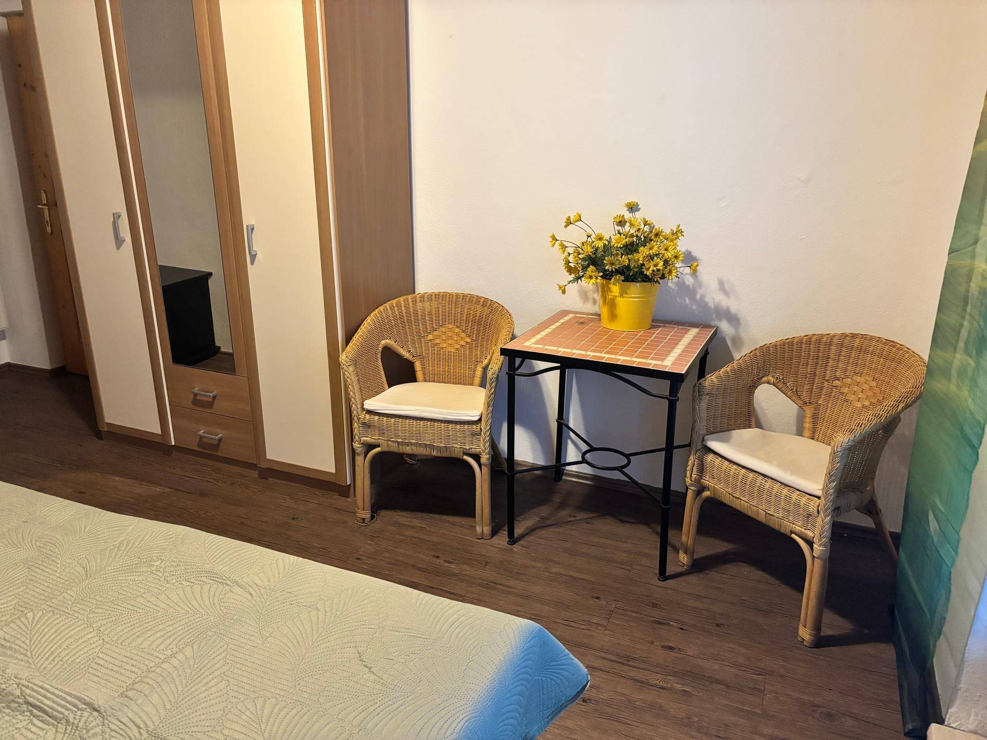 Schlafzimmer mit Kleiderschrank, Korbstühlen und einem kleinen Tisch mit Blumen.