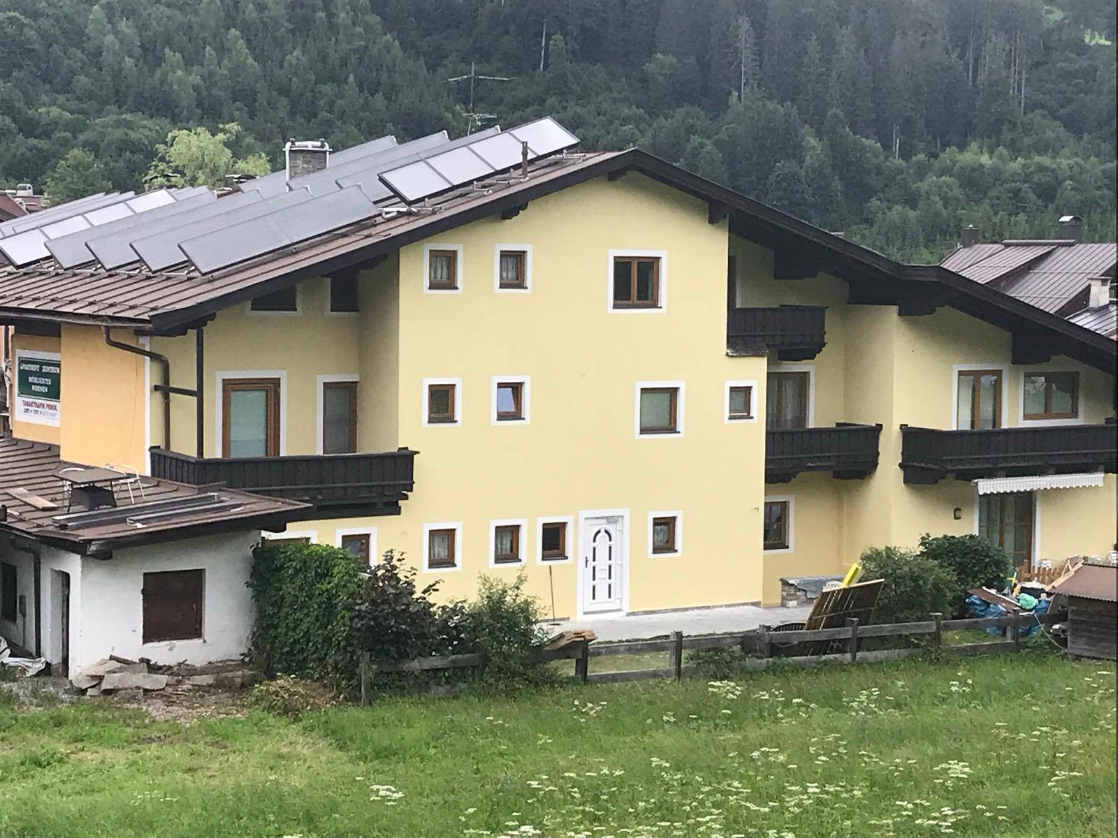 Gelbes Mehrfamilienhaus mit mehreren Balkonen und Solarpaneelen auf dem Dach vor einer grünen Bergkulisse.