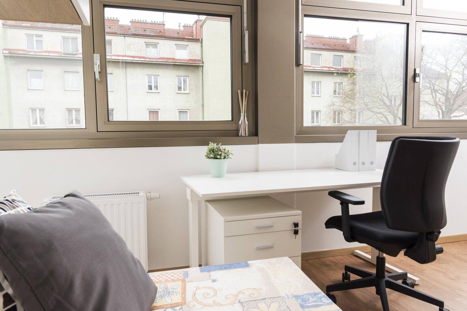 Arbeitsbereich mit Schreibtisch und Bürostuhl, hell durch große Fenster mit Stadtblick.