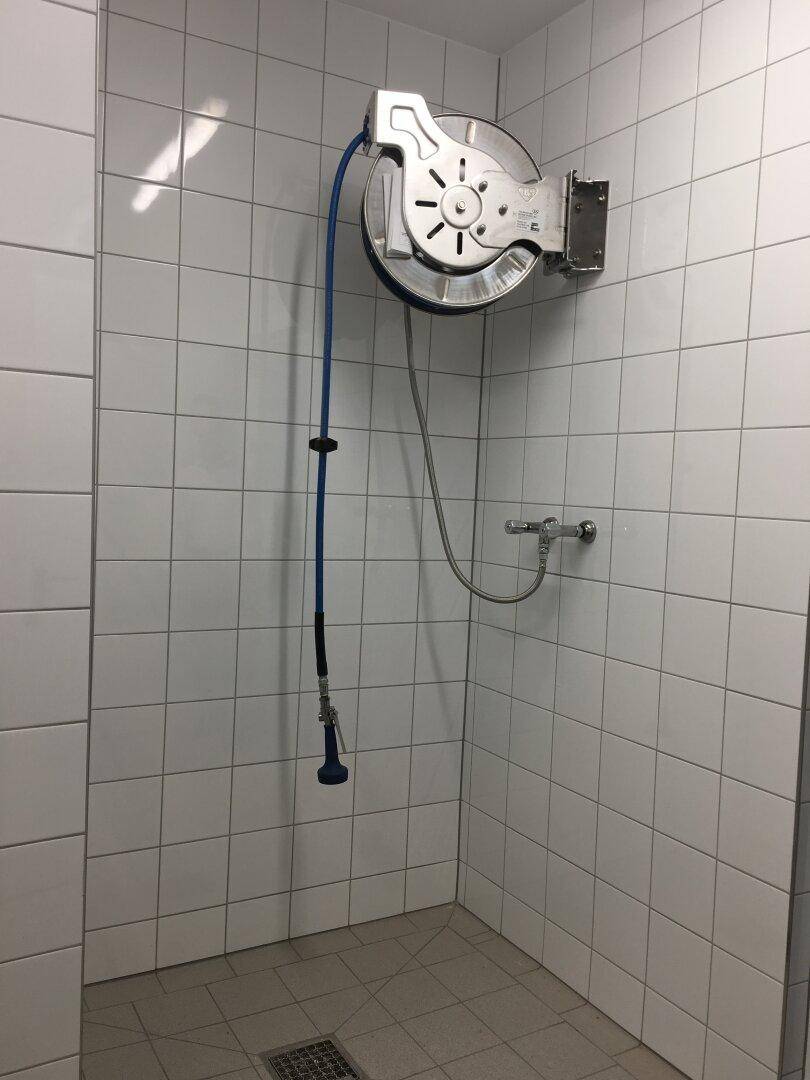 Gefliester Raum mit einer Wandhalterung für einen Schlauch und einem Wasseranschluss, möglicherweise ein Reinigungsbereich.