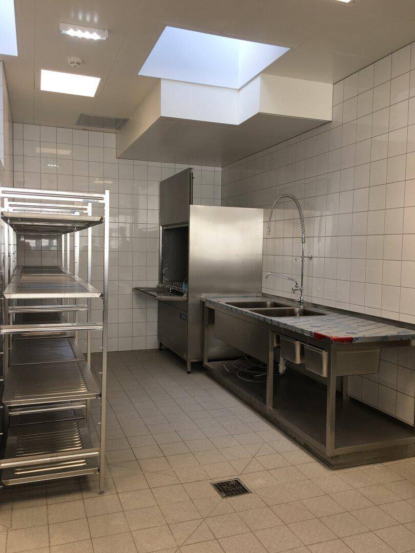 Industrielle Küche mit Edelstahlspüle, Geschirrspüler und Regalen, geflieste Wände und Deckenbeleuchtung.