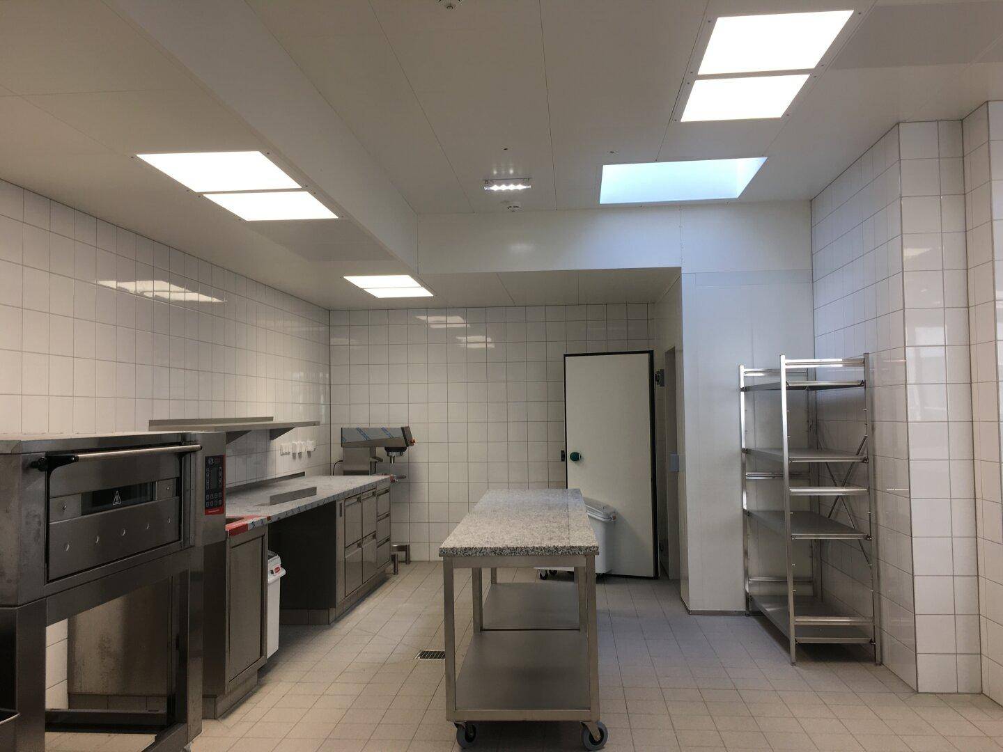 Professionelle Küche mit Edelstahlmöbeln, Backofen und Regalen, geflieste Wände und Deckenbeleuchtung.