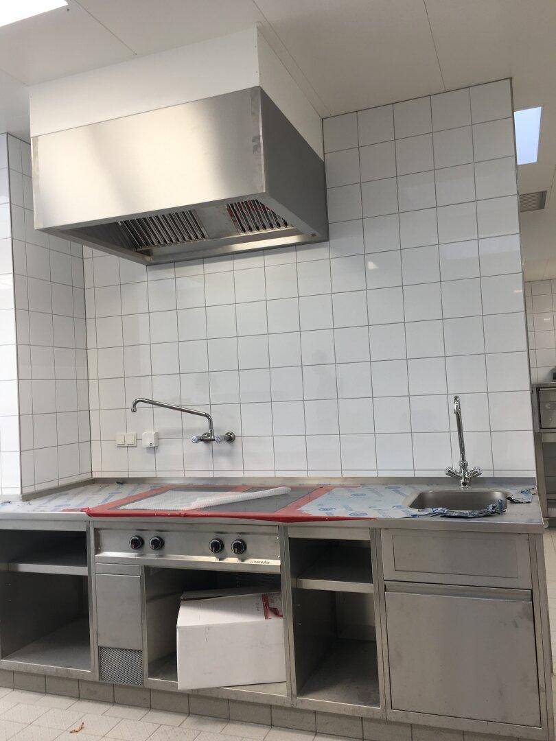 Küchenzeile aus Edelstahl mit Gaskochfeld, Spüle und Dunstabzugshaube, geflieste Wände.