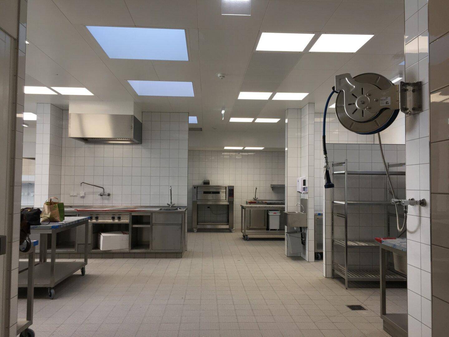 Große Industrieküche mit Edelstahlmöbeln, Spülbereich und Regalen, geflieste Wände und Deckenbeleuchtung.