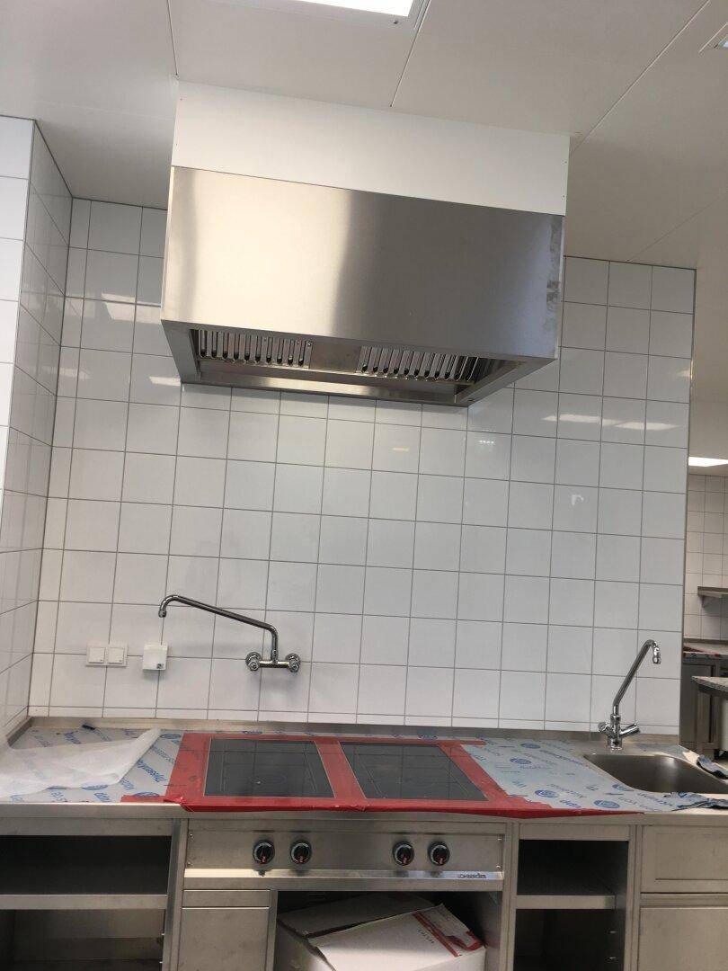 Küchenzeile aus Edelstahl mit Induktionskochfeldern, Spüle und Dunstabzugshaube, geflieste Wände.