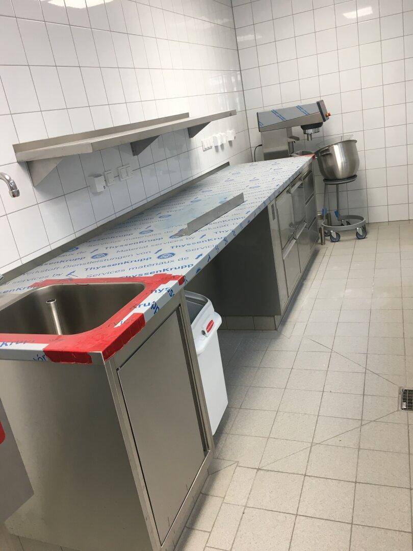 Lange Edelstahl-Arbeitsplatte mit integriertem Spülbecken und Wandregalen in einer gefliesten Gewerbeküche.