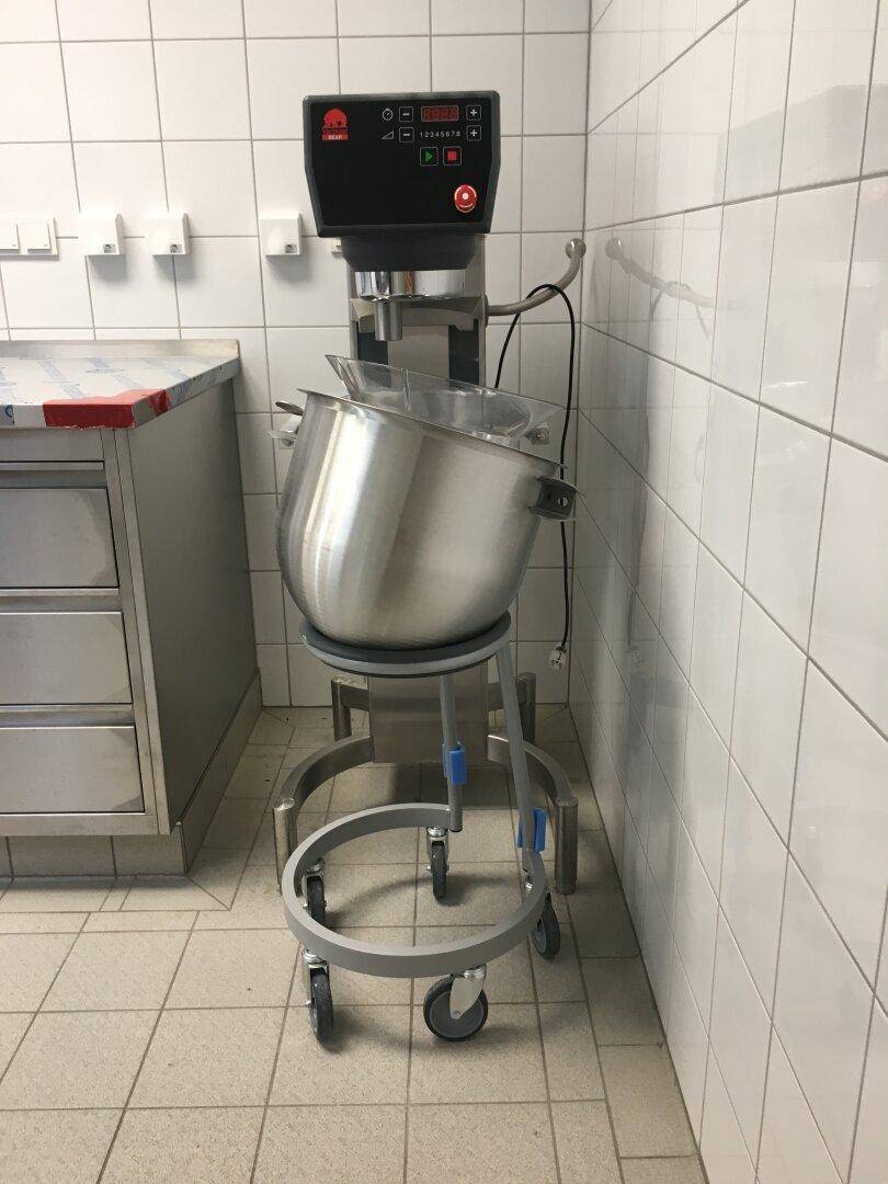 Professionelle Küchenmaschine mit großem Edelstahlkessel auf Rollen in einer gefliesten Gewerbeküche.