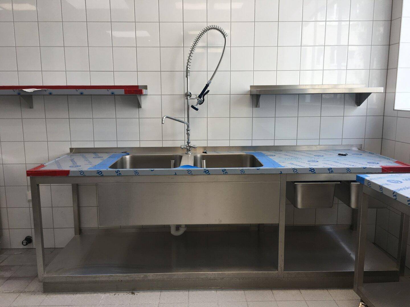 Große Doppelspüle aus Edelstahl mit professioneller Armatur und Wandregalen in einer gefliesten Gewerbeküche.