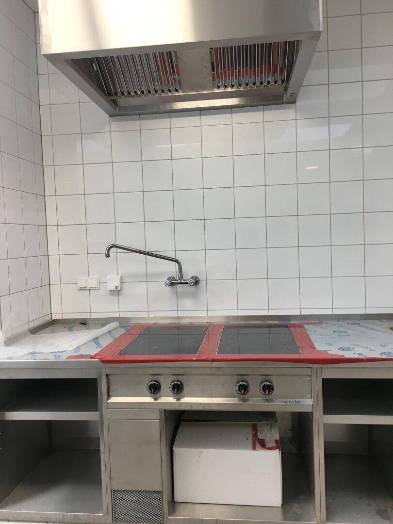 Professionelle Kochfelder mit Dunstabzugshaube und Edelstahl-Arbeitsplatte in einer gefliesten Gewerbeküche.