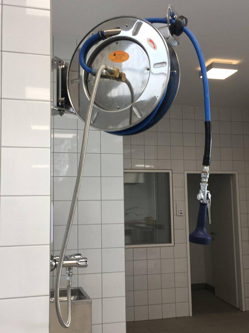 Wandmontierte Schlauchtrommel mit blauer Sprühdüse und Edelstahlarmatur in einer gefliesten Umgebung.