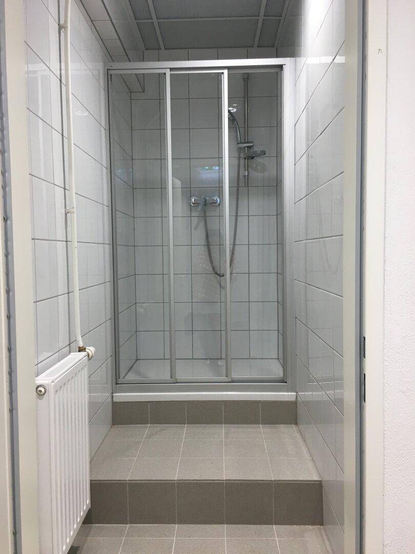 Duschkabine mit Glaswänden und Heizkörper in einem gefliesten Raum mit grauen Bodenfliesen.