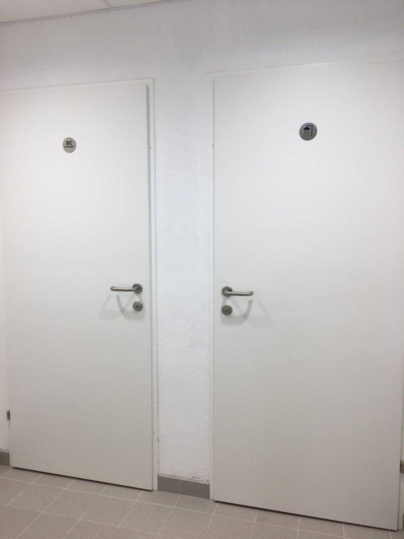 Zwei weiße Türen mit Piktogrammen für WC und Dusche in einem gefliesten Flur.
