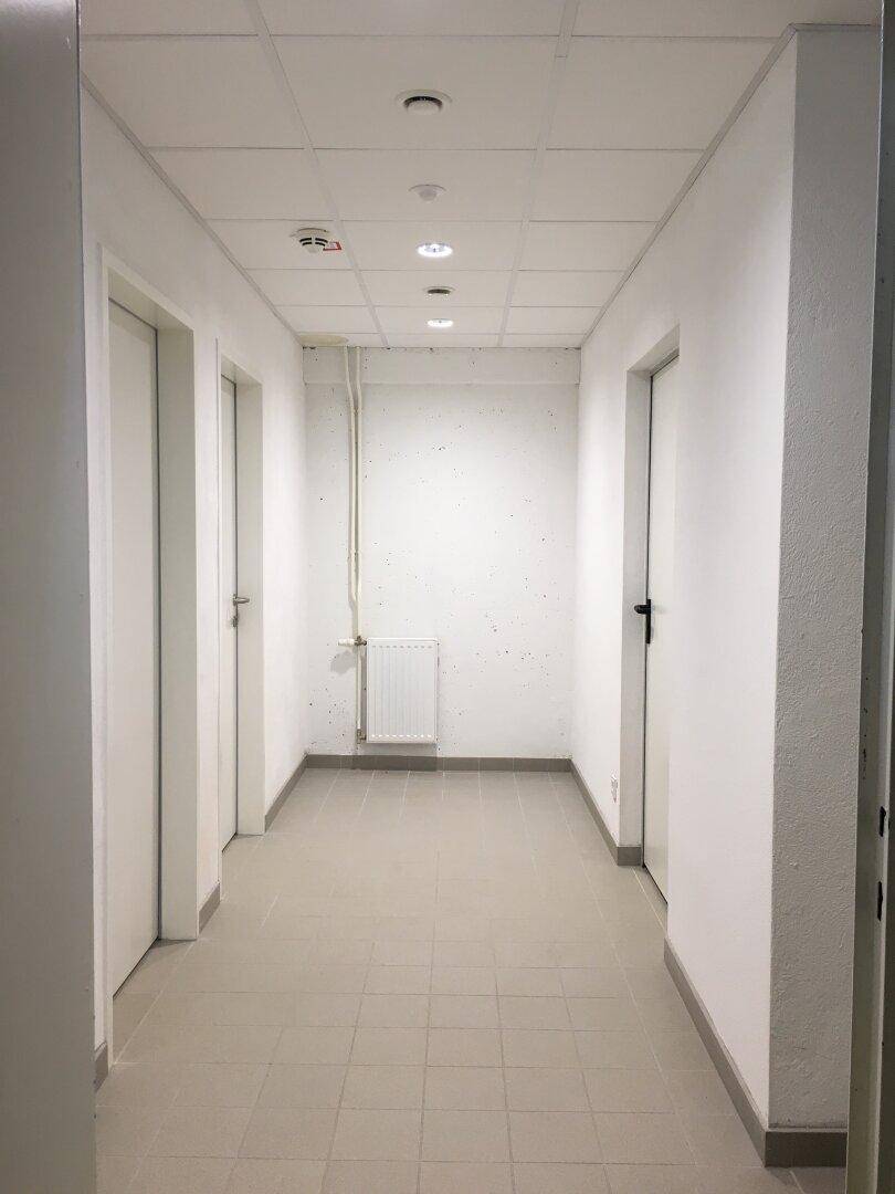 Langer, heller Flur mit weißen Wänden, grauen Bodenfliesen und Deckenbeleuchtung, führt zu mehreren Türen.