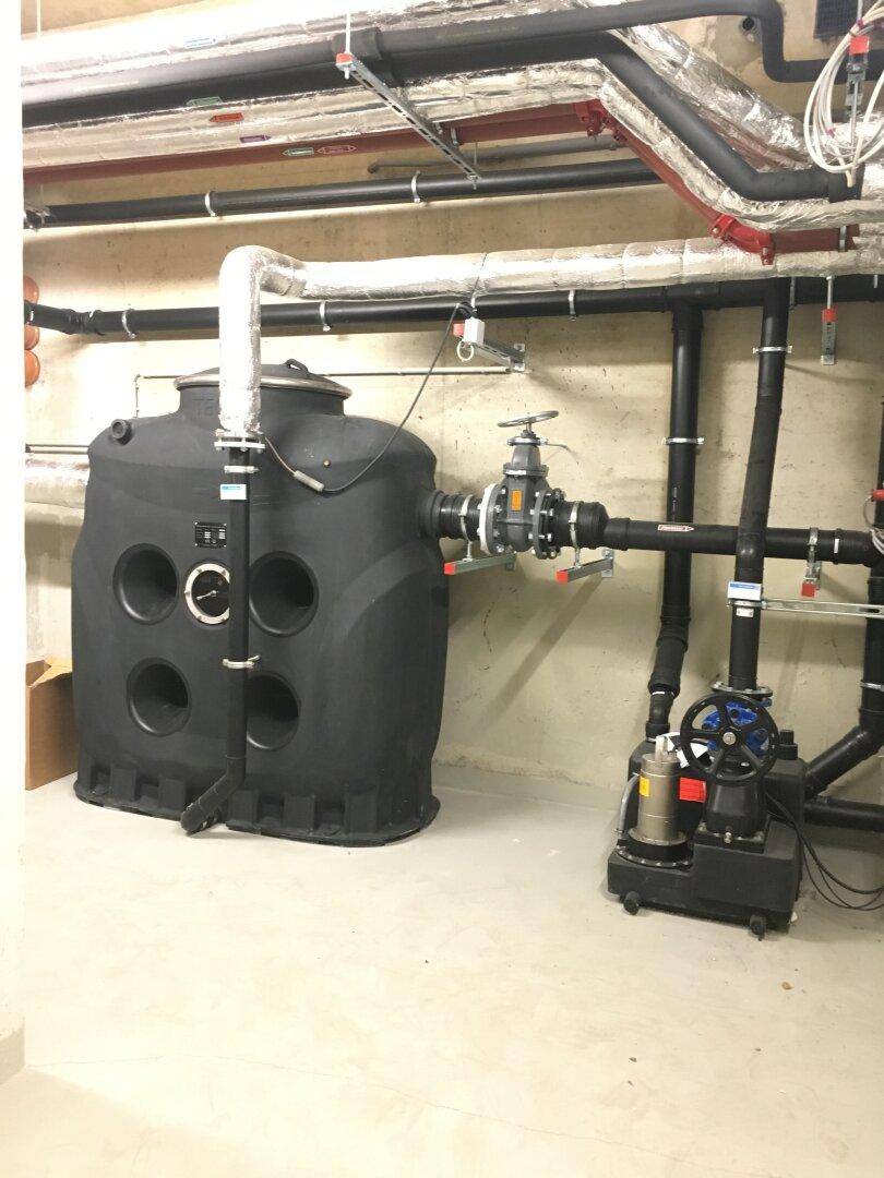 Technischer Raum mit großen Rohren, Ventilen und einem schwarzen Wassertank auf dem Boden.