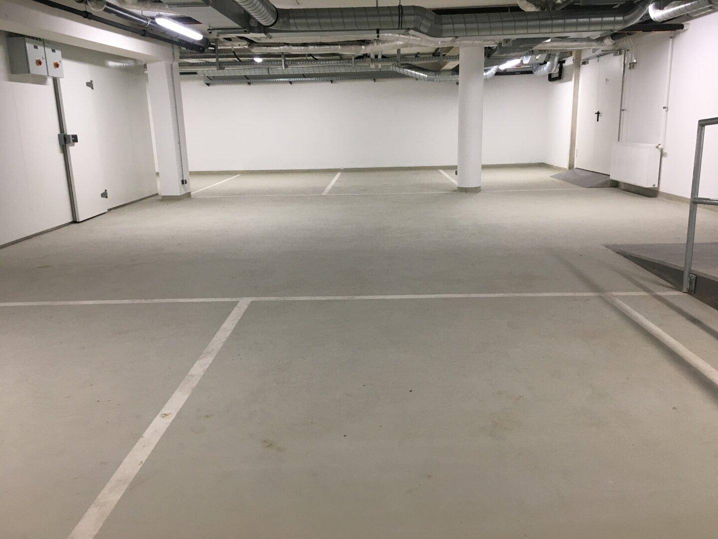 Geräumige Tiefgarage mit markierten Stellplätzen, weißen Wänden und sichtbaren Lüftungsrohren an der Decke.