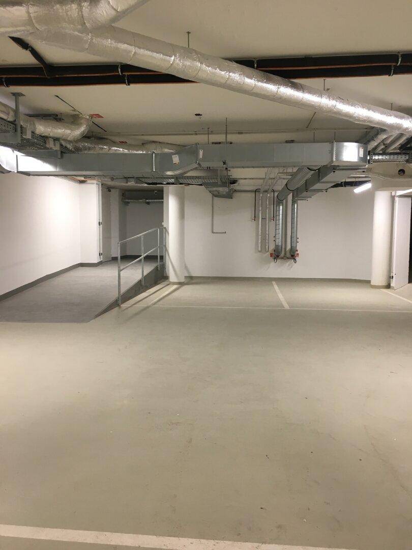 Weitläufige Tiefgarage mit einer Rampe, markierten Stellplätzen und sichtbaren Installationen an der Decke.