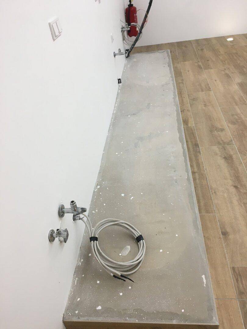 Lange Betonarbeitsplatte mit sichtbaren Anschlüssen und Kabeln in einem Raum mit Holzoptik-Fliesen.