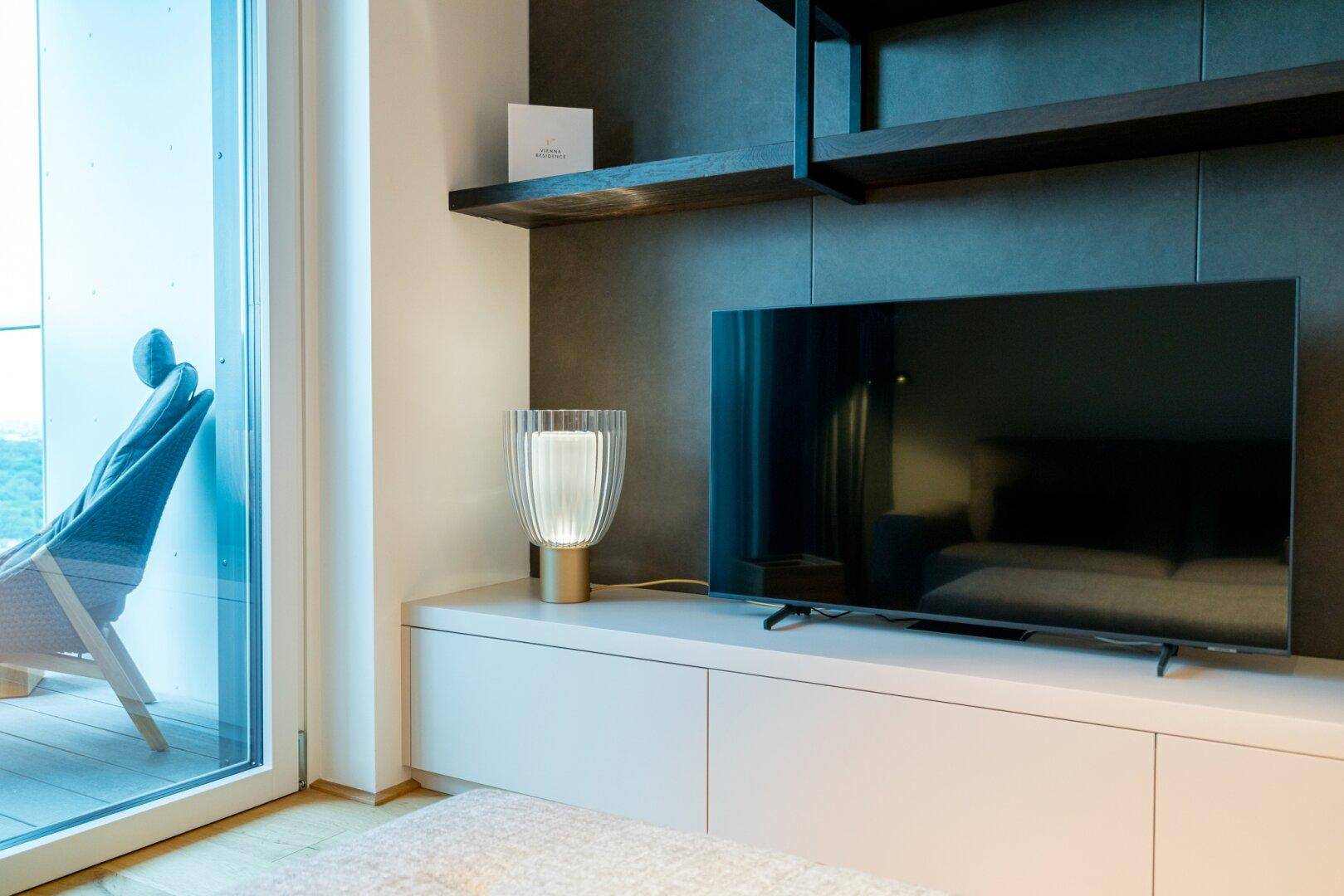 Heller Wohnbereich mit Fernseher und Zugang zu einem Balkon, der viel Tageslicht hereinlässt.