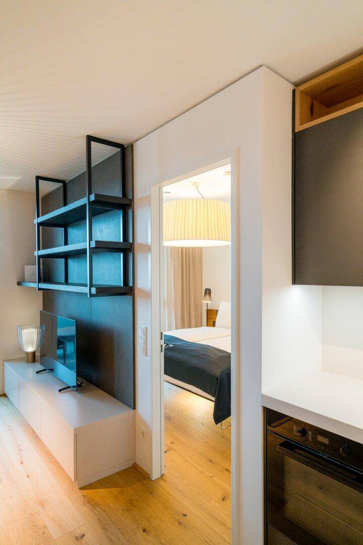 Wohnbereich mit Regalsystem und Blick in ein helles Schlafzimmer mit großem Bett.