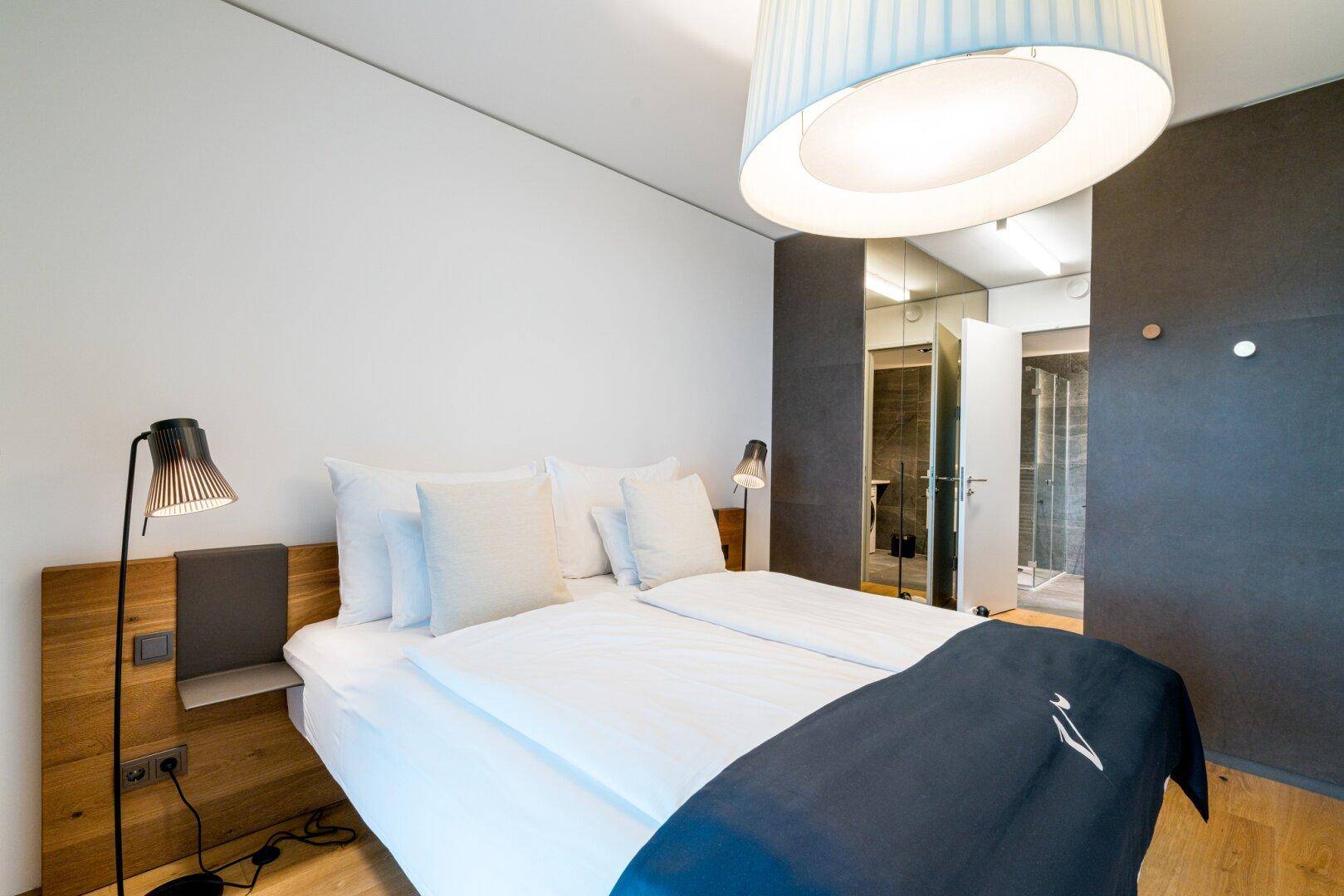 Modernes Schlafzimmer mit Doppelbett, Holzkopfteil und direktem Zugang zum Badezimmer.
