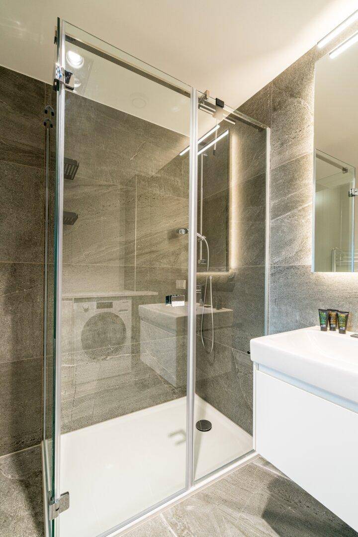 Elegantes Badezimmer mit großer Glasdusche, modernem Waschtisch und grauen Fliesen.