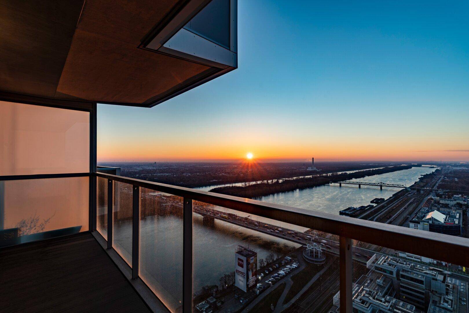Balkon mit Glasbrüstung und weitem Blick über den Fluss und die Stadt bei Sonnenuntergang.