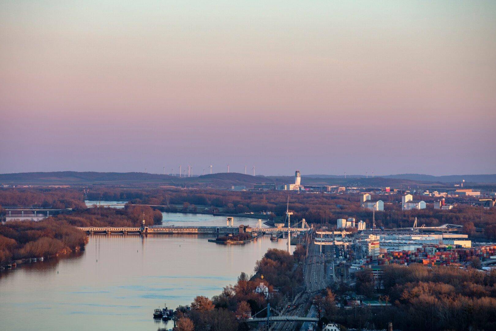 Panoramablick auf den Fluss mit Brücken, Industrieanlagen und bewaldeten Ufern in der Abenddämmerung.