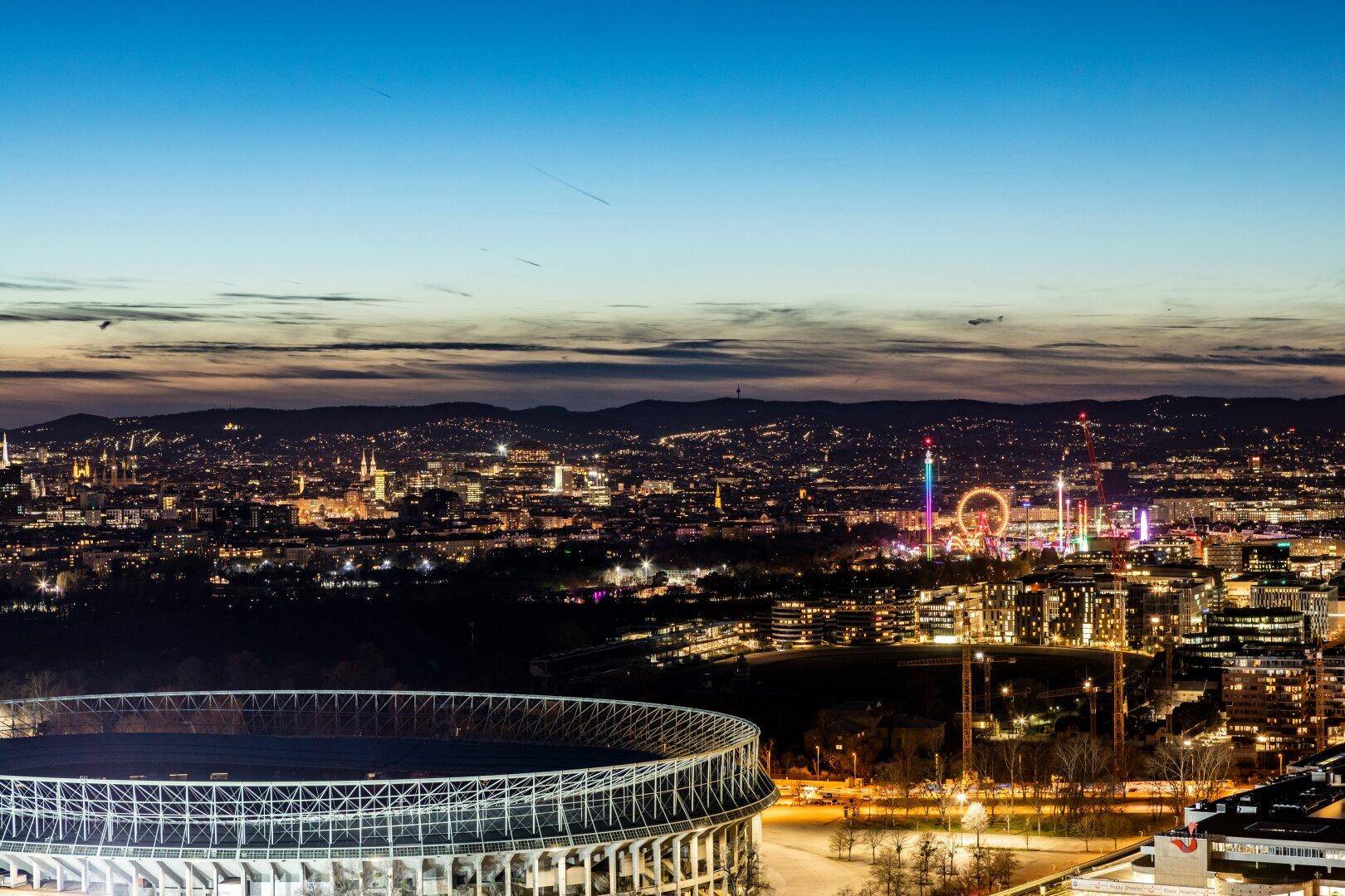 Nachtansicht der Stadt mit einem beleuchteten Stadion und dem Prater im Hintergrund.
