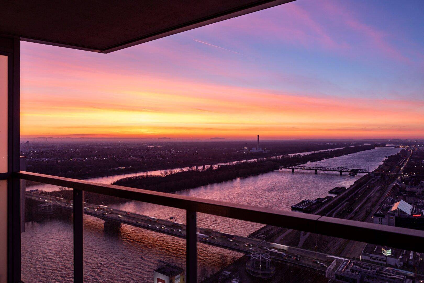 Balkon mit Glasbrüstung und weitem Blick über den Fluss und die Stadt bei Sonnenuntergang.
