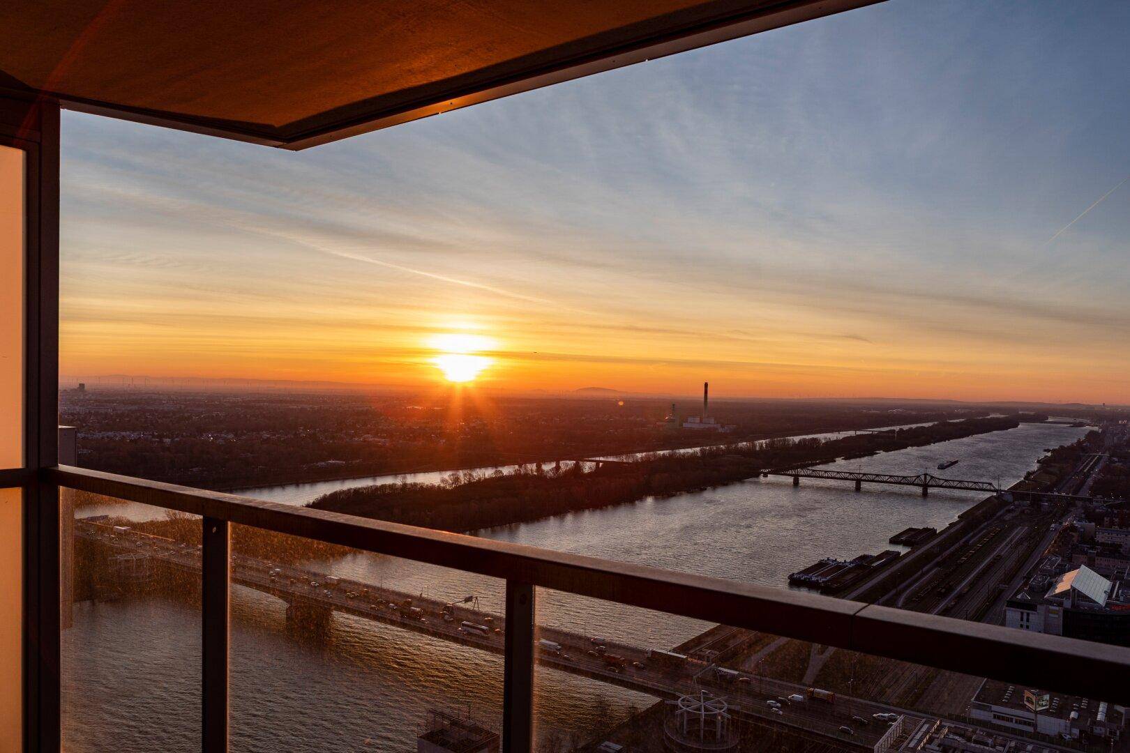 Balkon mit Glasbrüstung und weitem Blick über den Fluss und die Stadt bei Sonnenuntergang.