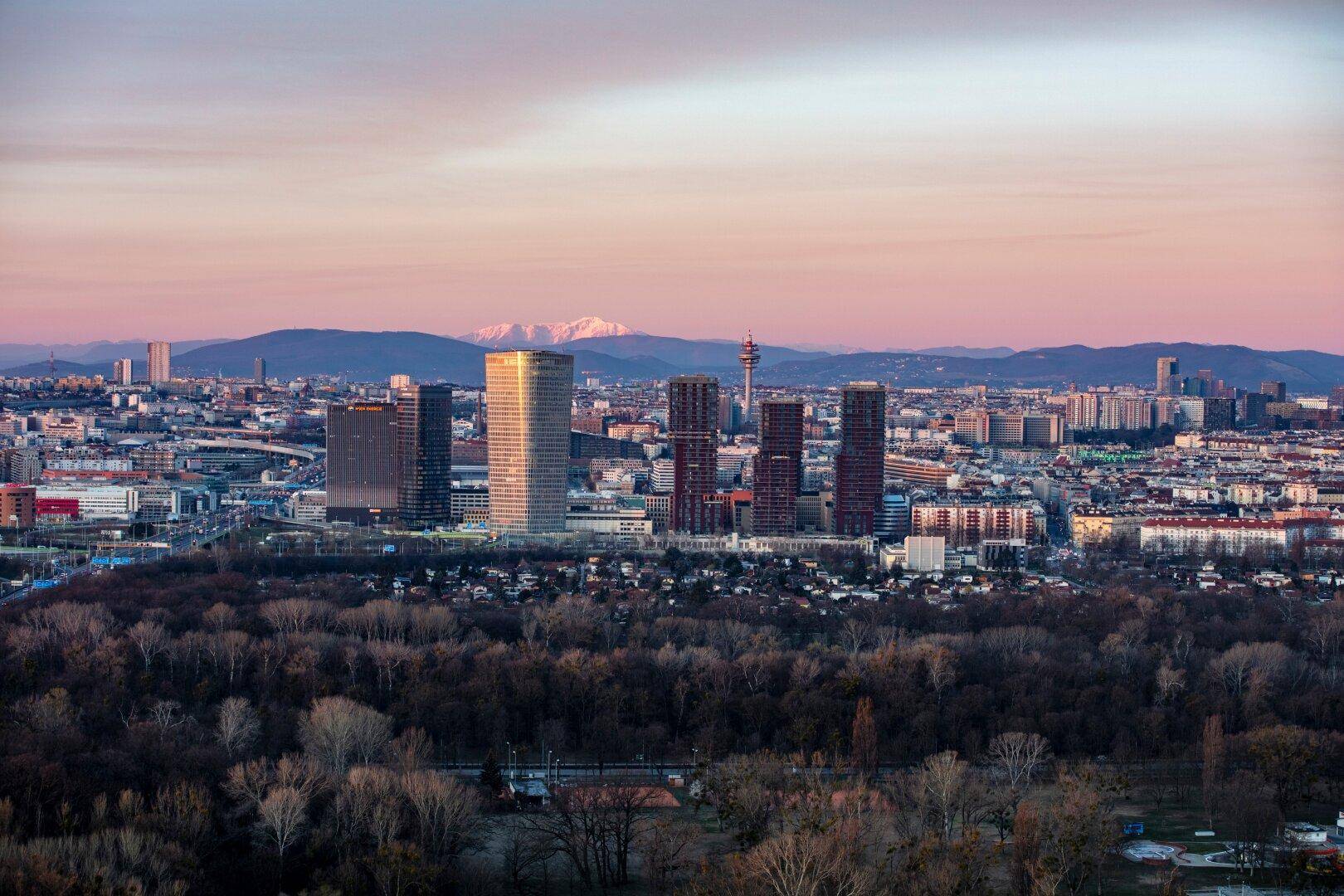 Panoramablick über die Stadt mit Hochhäusern, Wäldern und schneebedeckten Bergen im Hintergrund bei Sonnenuntergang.