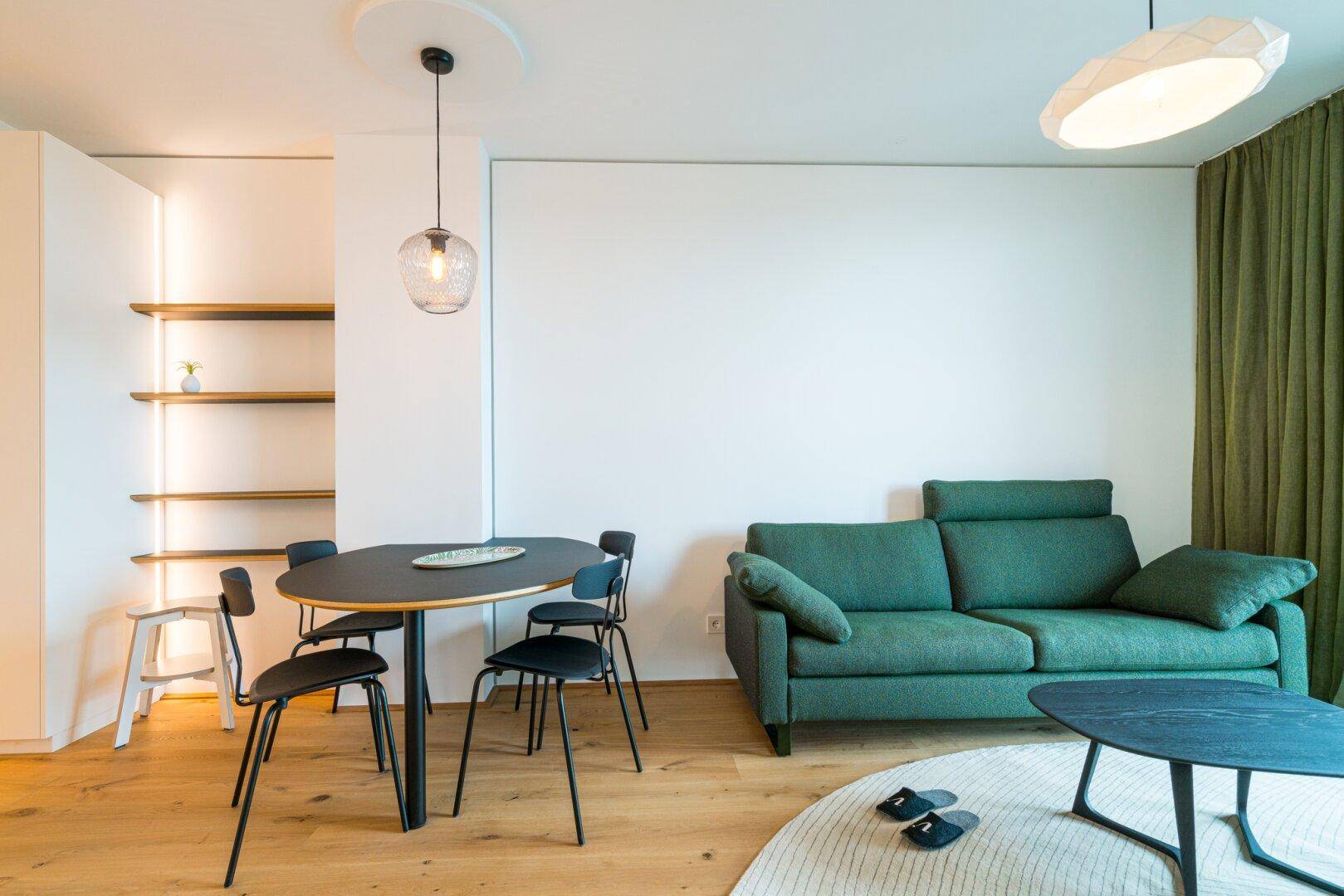 Heller Wohn- und Essbereich mit grünem Sofa, Holzboden und modernen Pendelleuchten.
