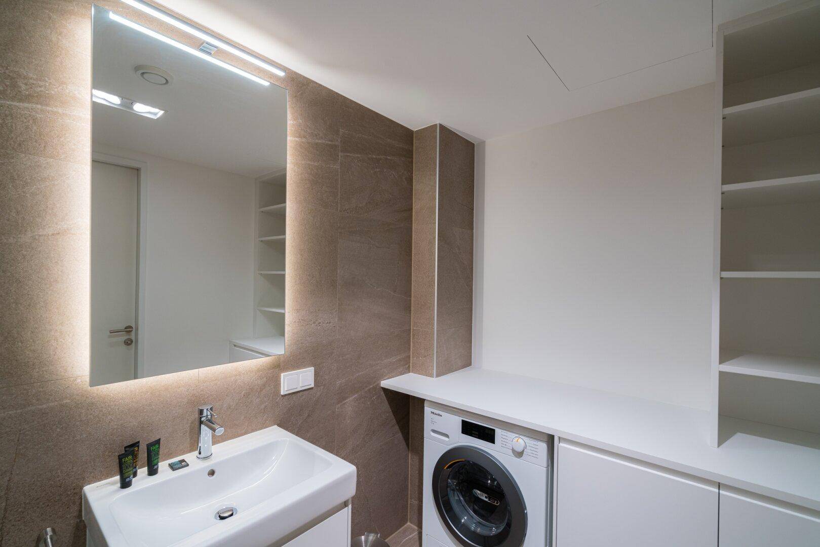 Modernes Badezimmer mit Waschbecken, beleuchtetem Spiegel und integrierter Waschmaschine.