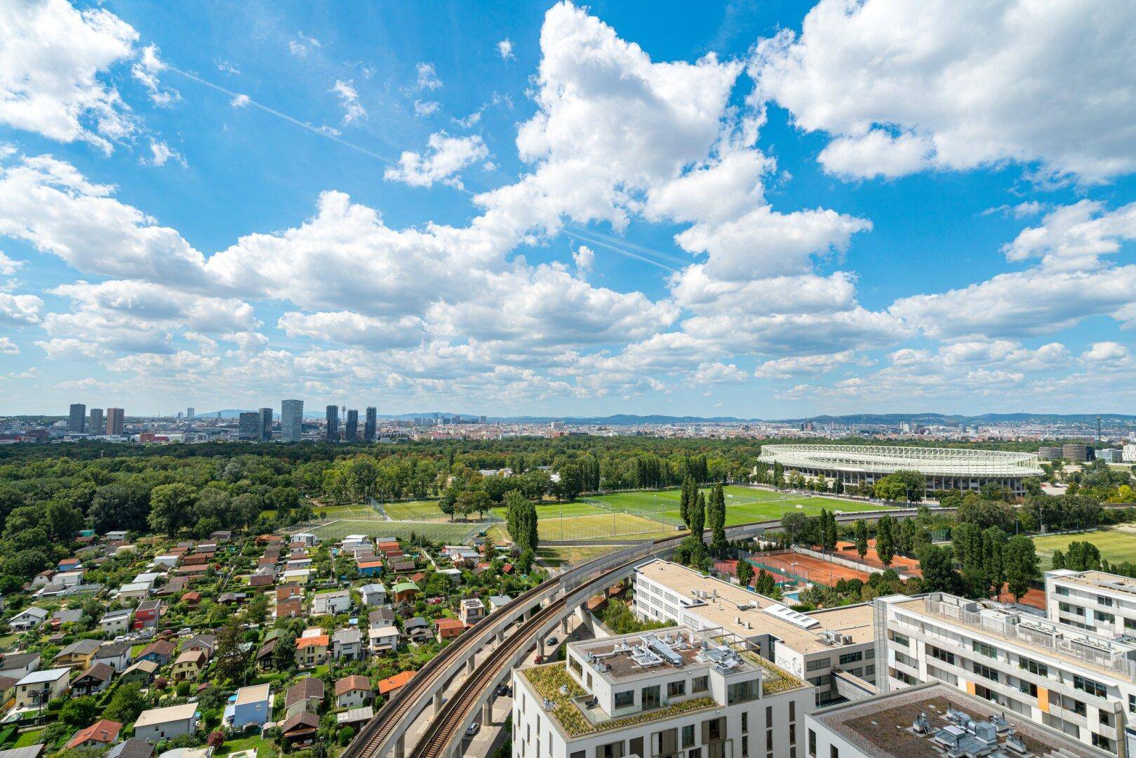 Weitläufiger Blick über die Stadt, grüne Parks und Sportanlagen unter blauem Himmel.