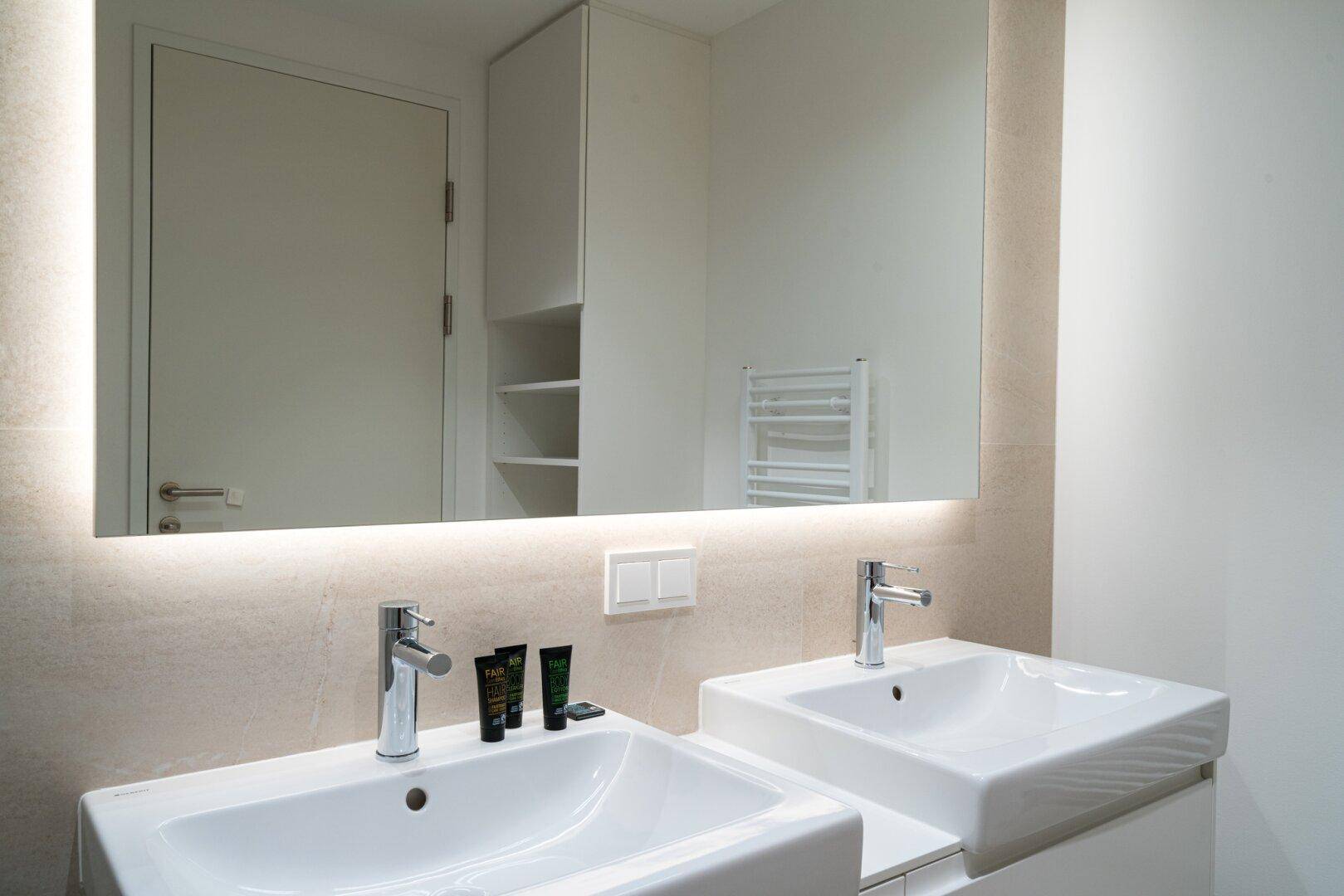 Modernes Badezimmer mit Doppelwaschbecken, beleuchtetem Spiegel und hellen Fliesen.