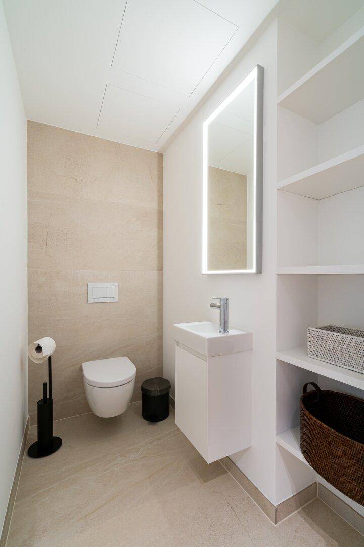 Modernes Gäste-WC mit wandhängendem WC, kleinem Waschbecken und beleuchtetem Spiegel.