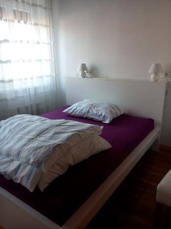 Helles Schlafzimmer mit Doppelbett, lila Matratze und gestreifter Bettwäsche, mit viel Tageslicht.