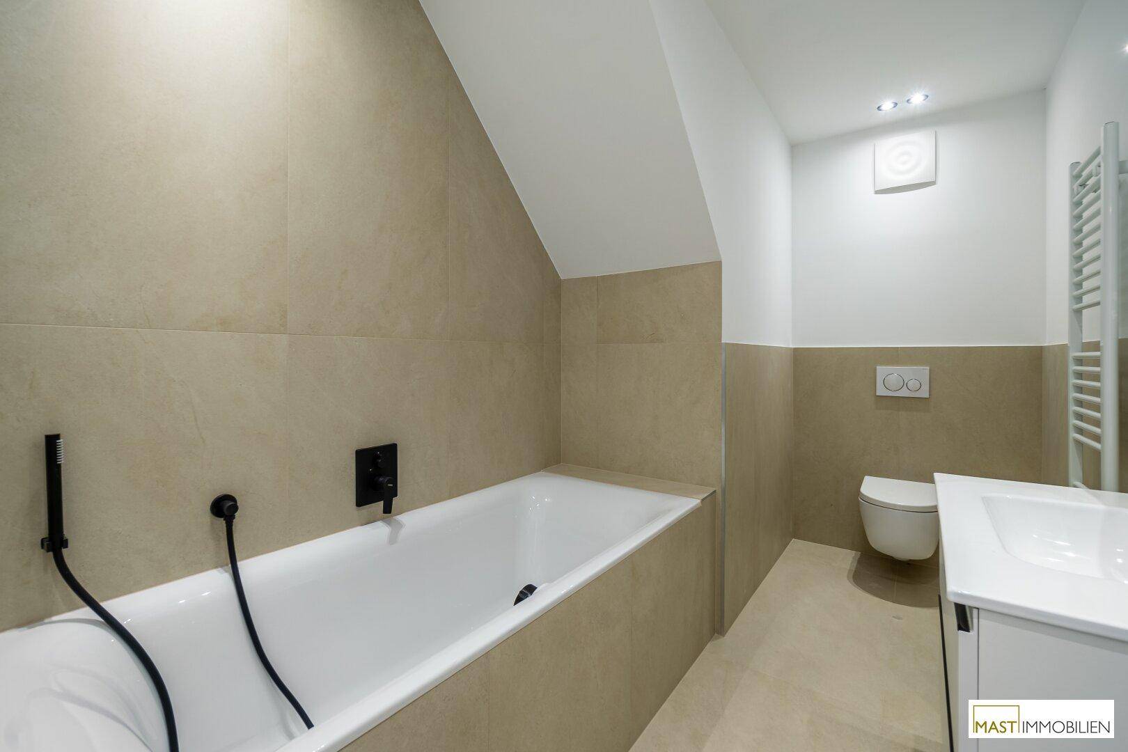 Modernes Badezimmer mit Badewanne, beigefarbenen Fliesen und Wand-WC unter der Dachschräge.