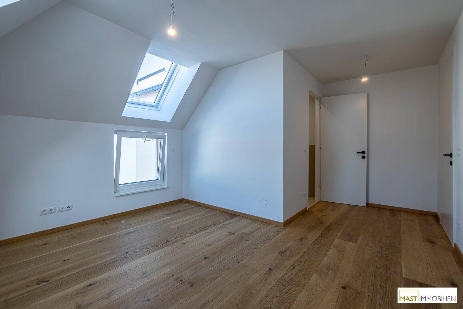 Helles Zimmer mit Holzboden, Dachschräge und einem Fenster, das viel Tageslicht hereinlässt.