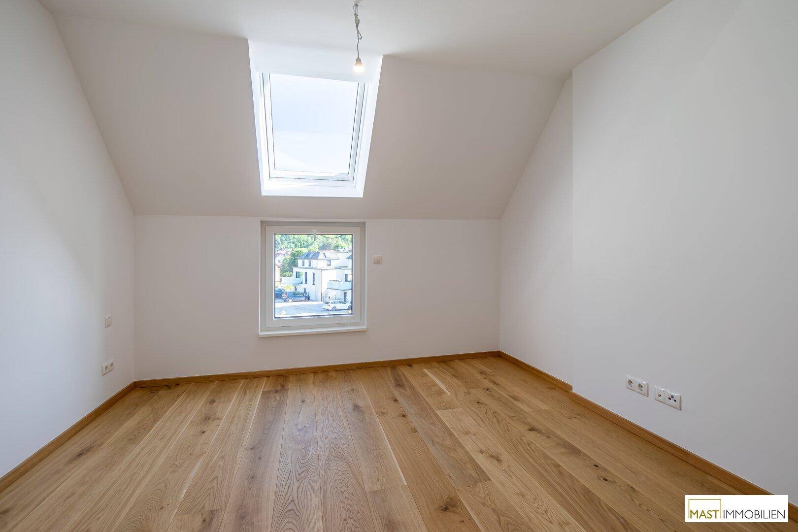 Helles Zimmer mit Holzboden, Dachschräge und einem Fenster mit Blick nach draußen.