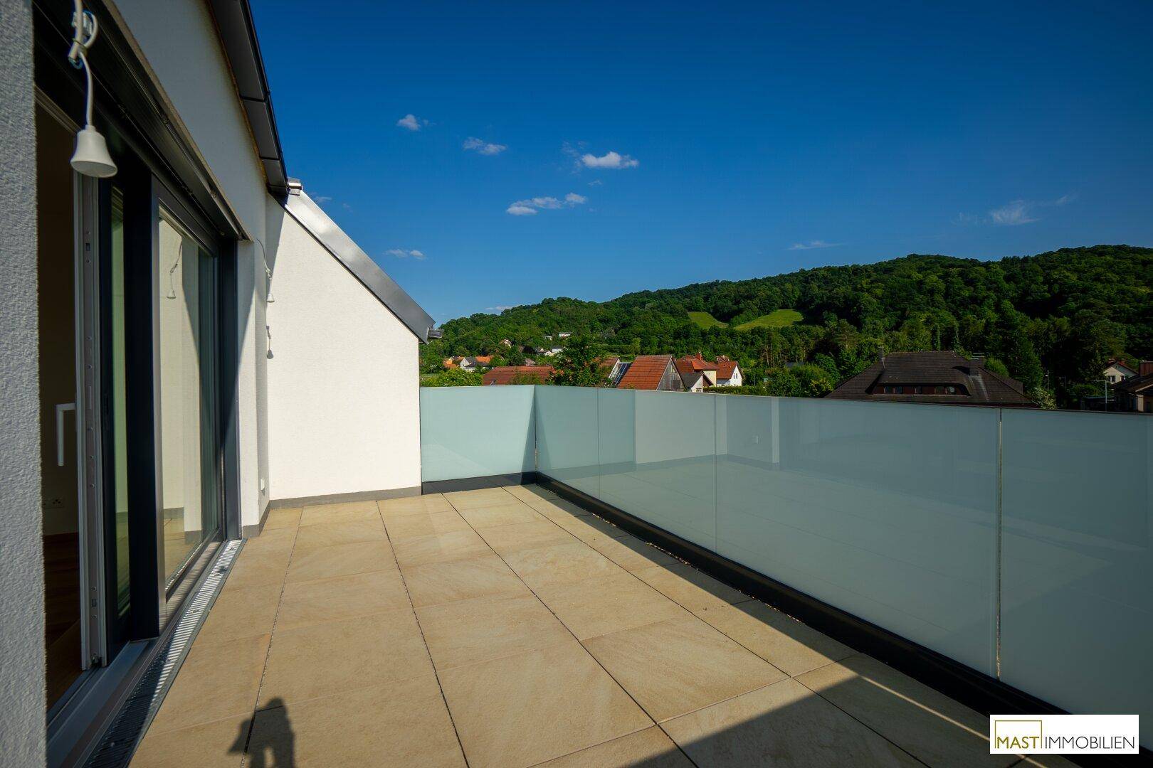 Großer Balkon mit Fliesenboden, Glasgeländer und Panoramablick auf die umliegenden Hügel.