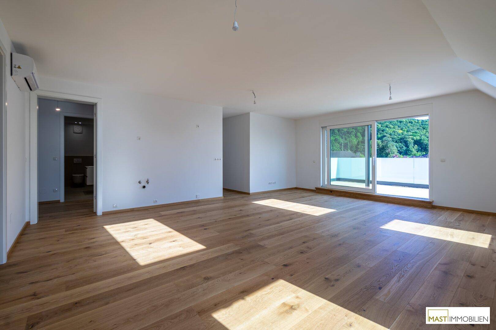 Geräumiges Zimmer mit Holzboden, Schiebetür zum Balkon und Blick auf die grüne Umgebung.