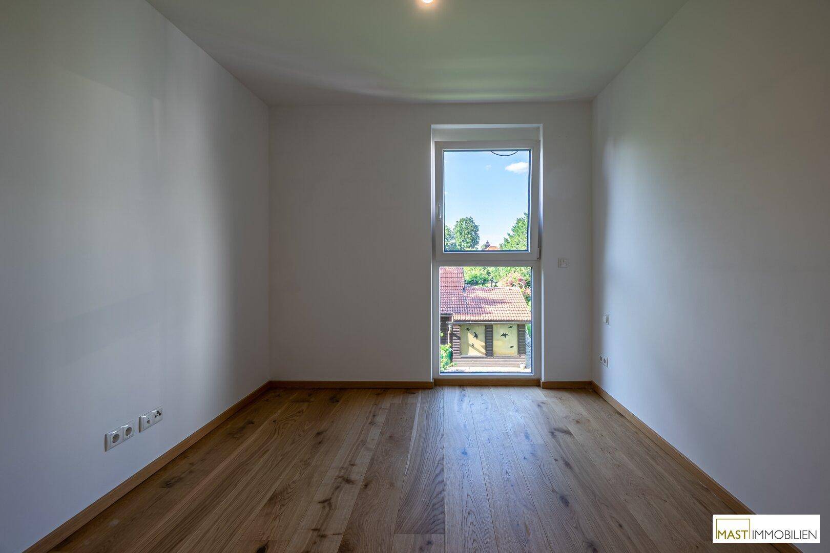 Helles Schlafzimmer mit Holzboden und einem Fenster mit Blick ins Grüne.