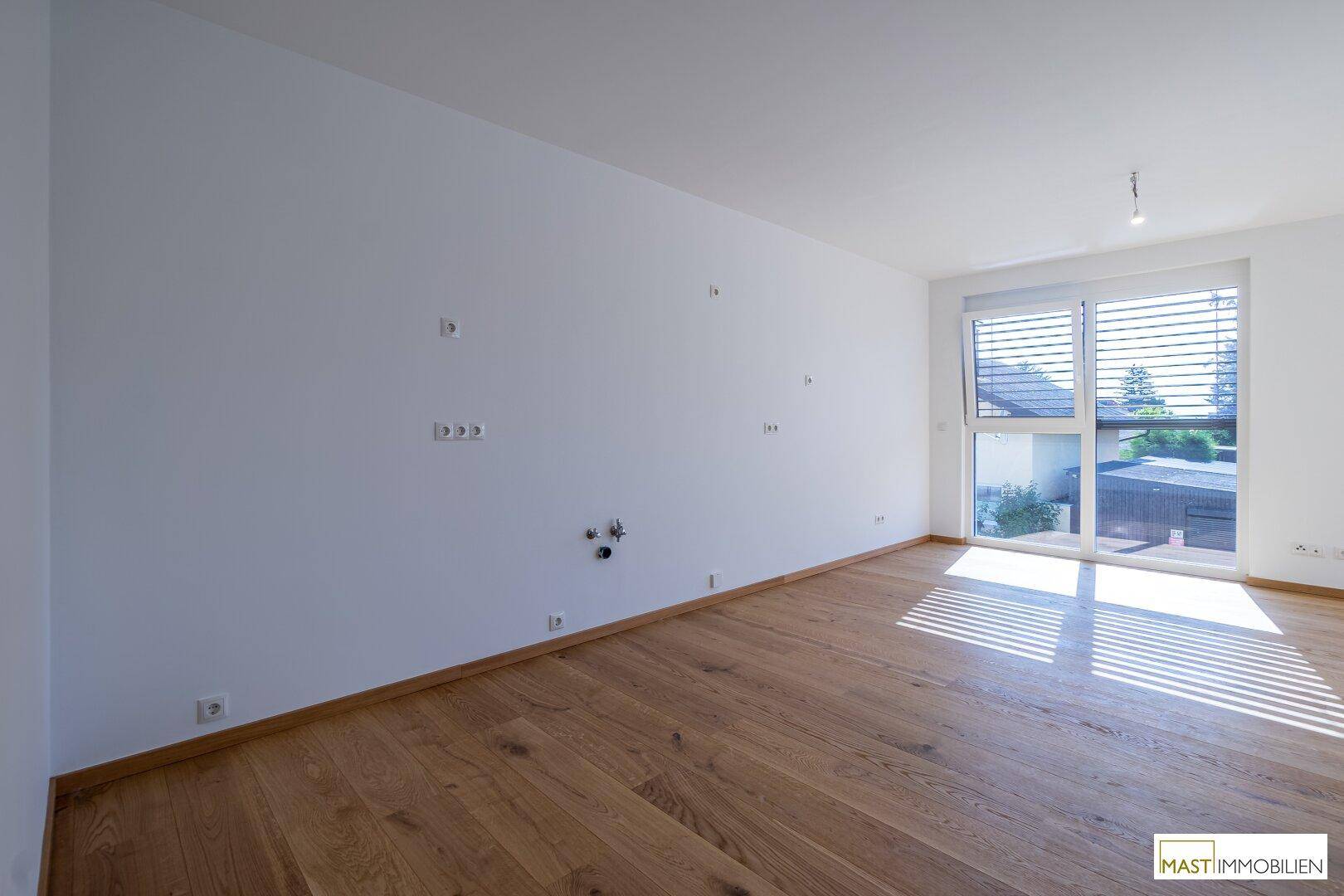 Unmöblierter Wohnbereich mit Holzboden und einem großen Fenster mit Außenjalousien.