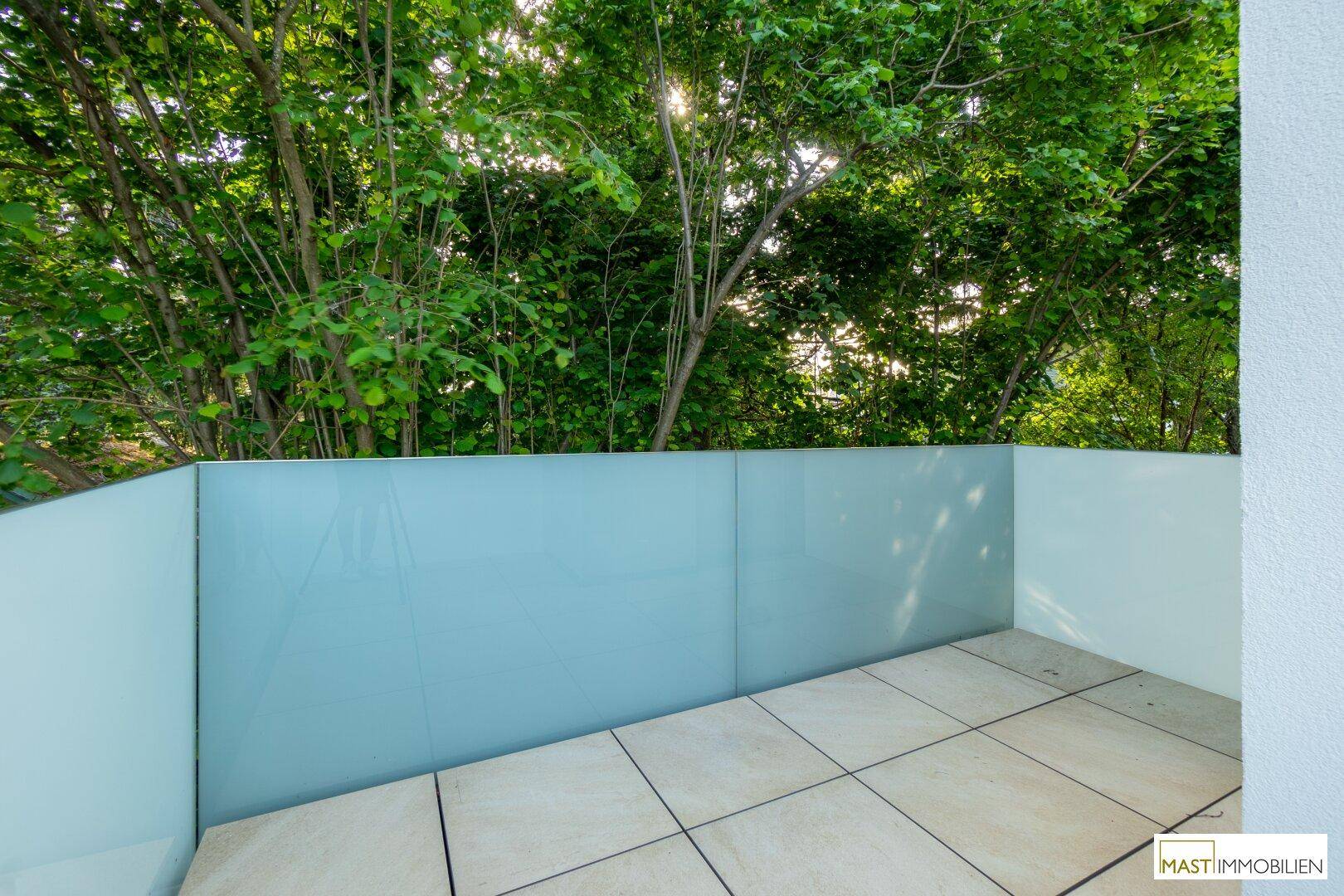 Heller Balkon mit Fliesenboden und blickdichter Brüstung, umgeben von grünen Bäumen.
