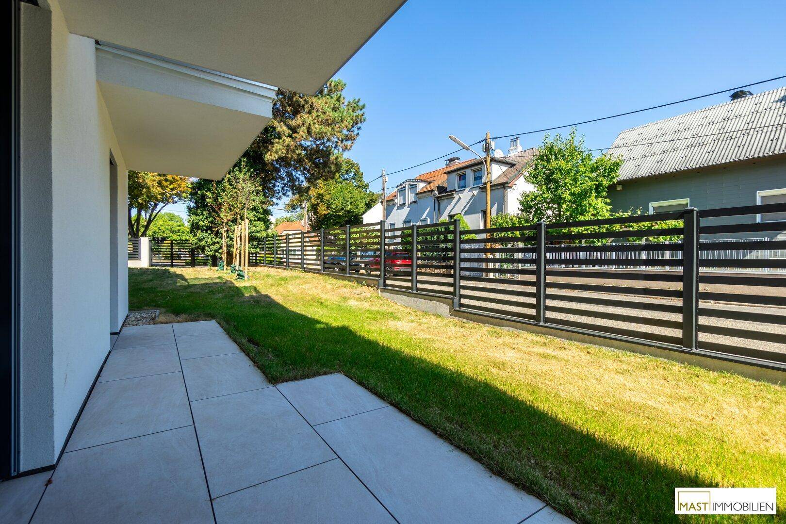 Sonniger Terrassenbereich mit Fliesenboden und angrenzender Rasenfläche, umgeben von einem modernen Zaun.