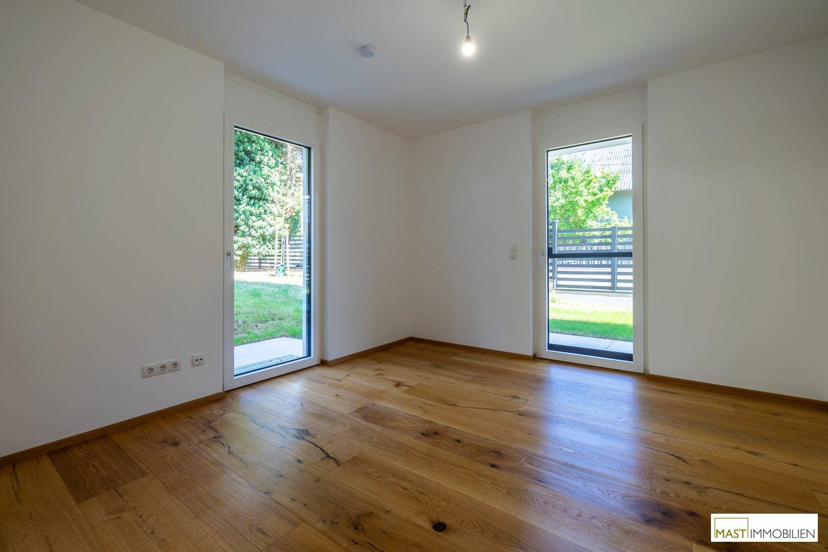 Helles Zimmer mit Holzfußboden und zwei hohen Fenstertüren, die den Blick ins Grüne freigeben.